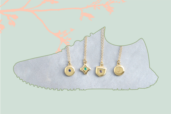 Outline of an Allbirds running shoe shown with four yellow gold birthstone and initial pendants, against a pale green backdrop with the pink outline of a branch.